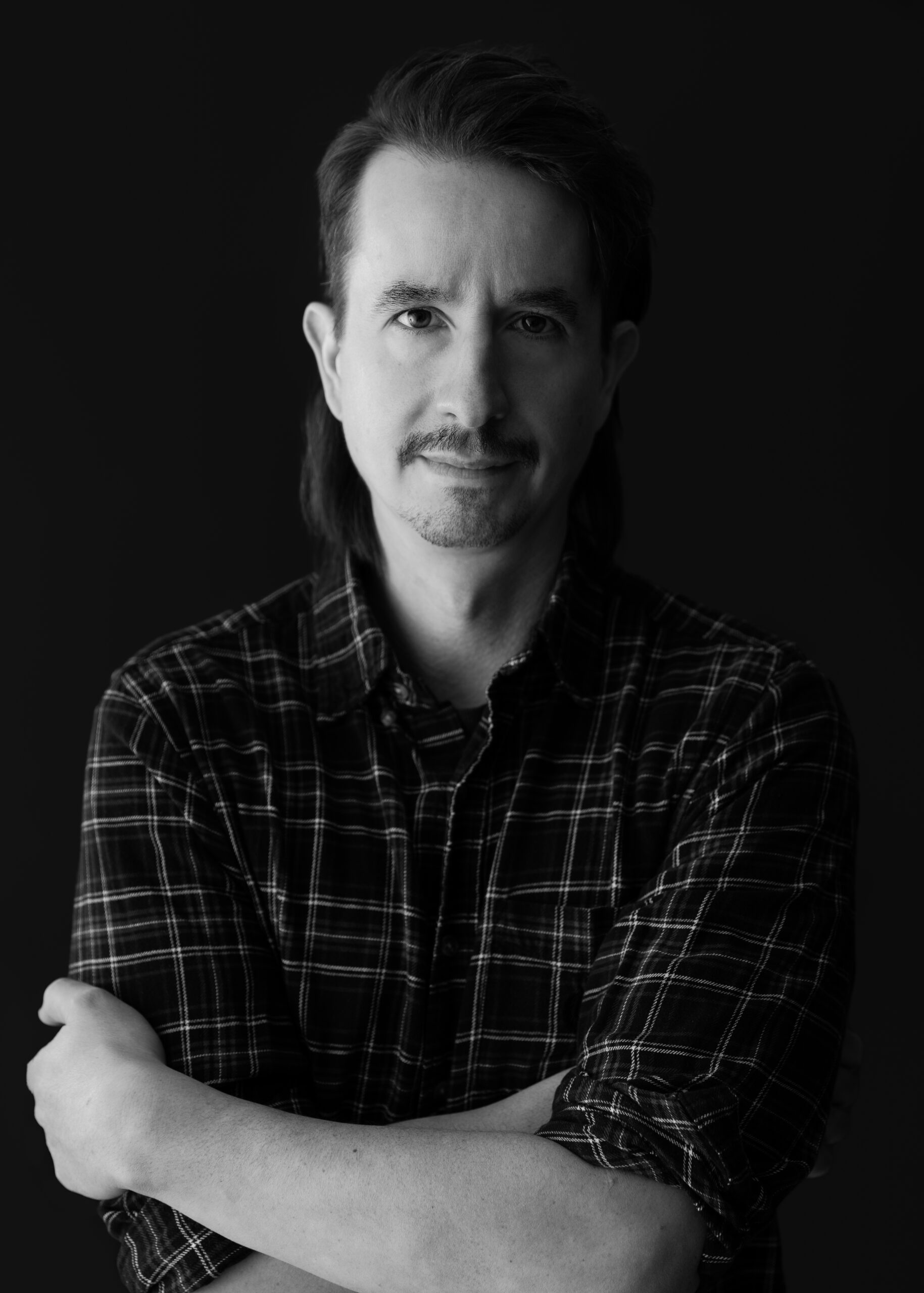 Black-and-white portrait of Spencer Gordon. He has light skin tone and dark eyes, hair, and eyebrows. He is wearing a plaid collared shirt. His bare left arm is crossed over his right.