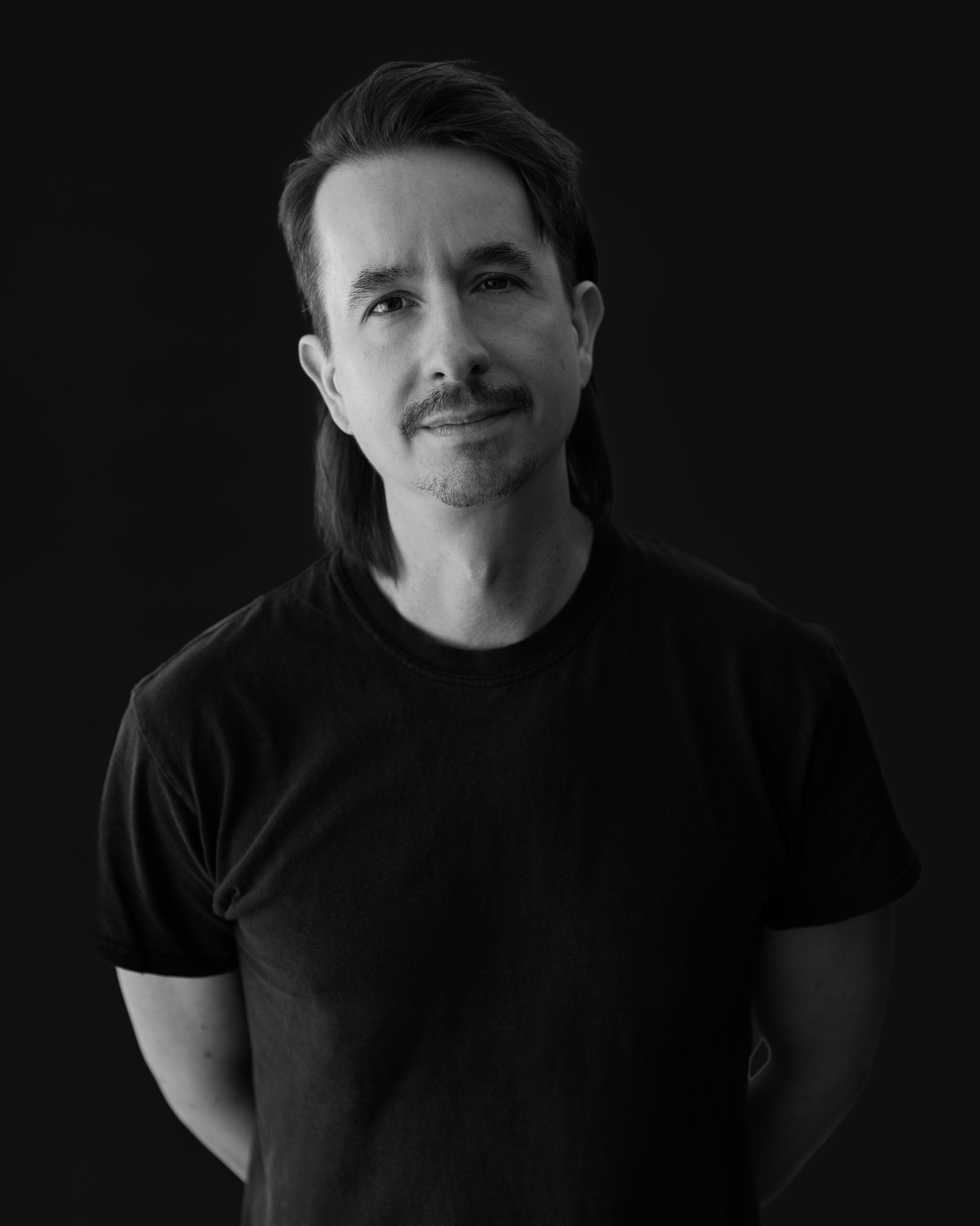 Black-and-white portrait of Spencer Gordon. He has light skin tone and dark eyes, hair, and eyebrows. He is wearing a dark t-shirt. His arms are held behind his back.